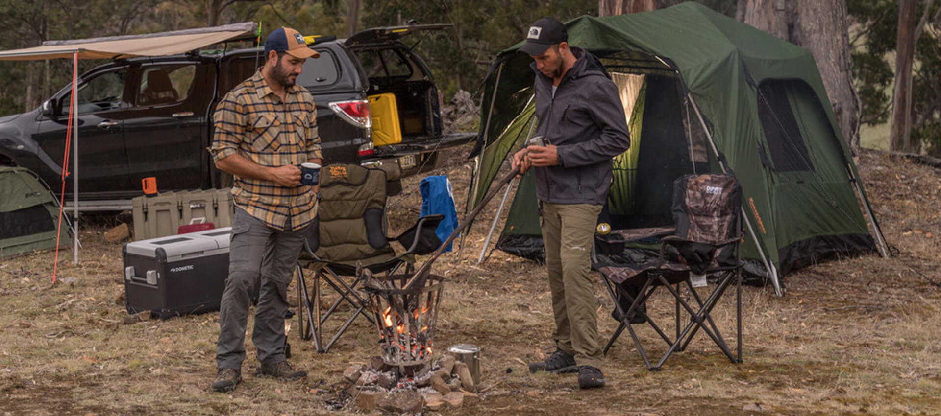 2 men standing around a campfire with pitched tent, camping chairs and portable fridges in the background